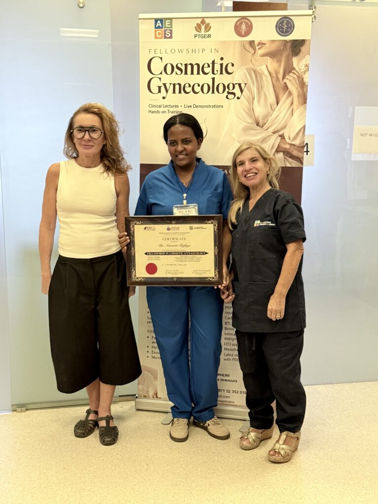 A woman in blue scrubs displays her certificate beside two women.