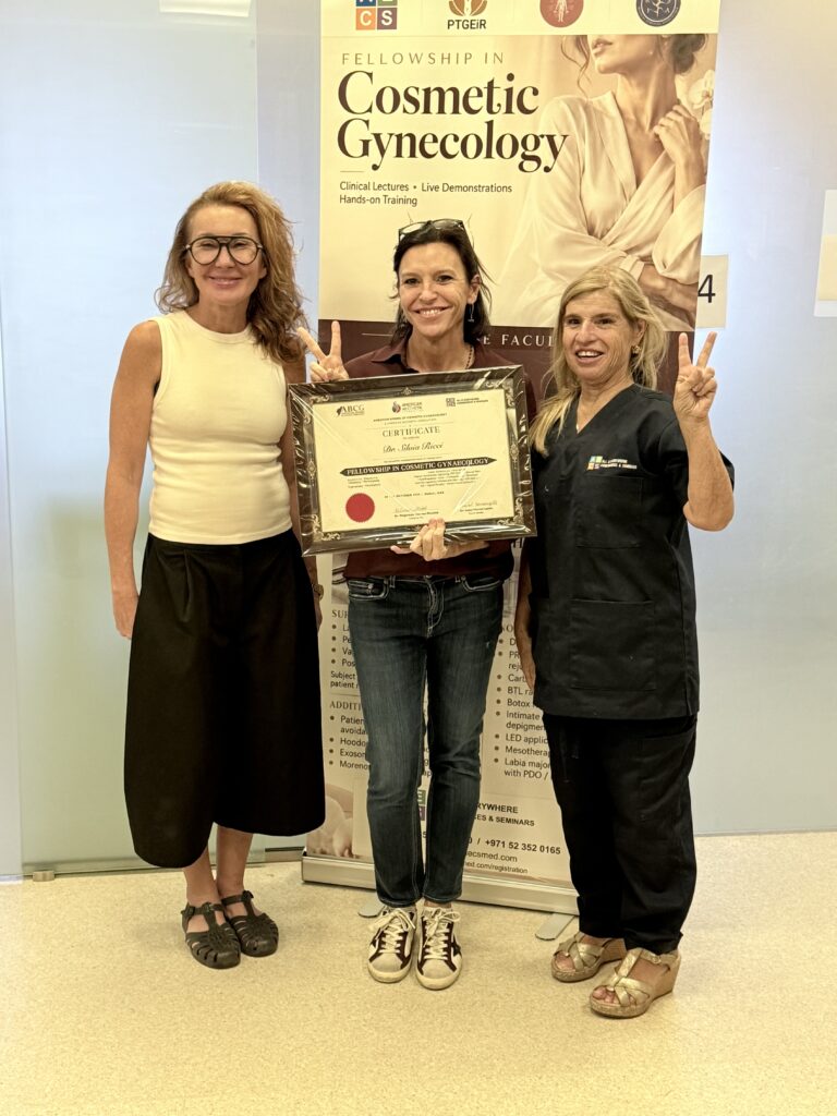 A woman in a dark red top and jeans displays her certificate beside two women.