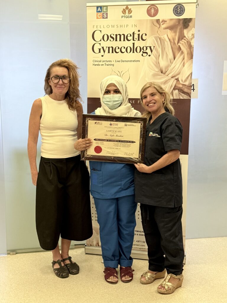 A masked woman in blue scrubs and a cream headscarf displays her certificate.